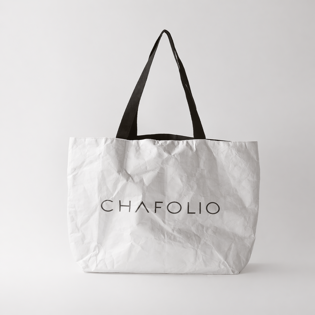 The Chafolio Tyvek Tote Bag is white with black handles and features "CHAFOLIO" printed in bold black letters on the front. The bag has a slightly crumpled appearance.