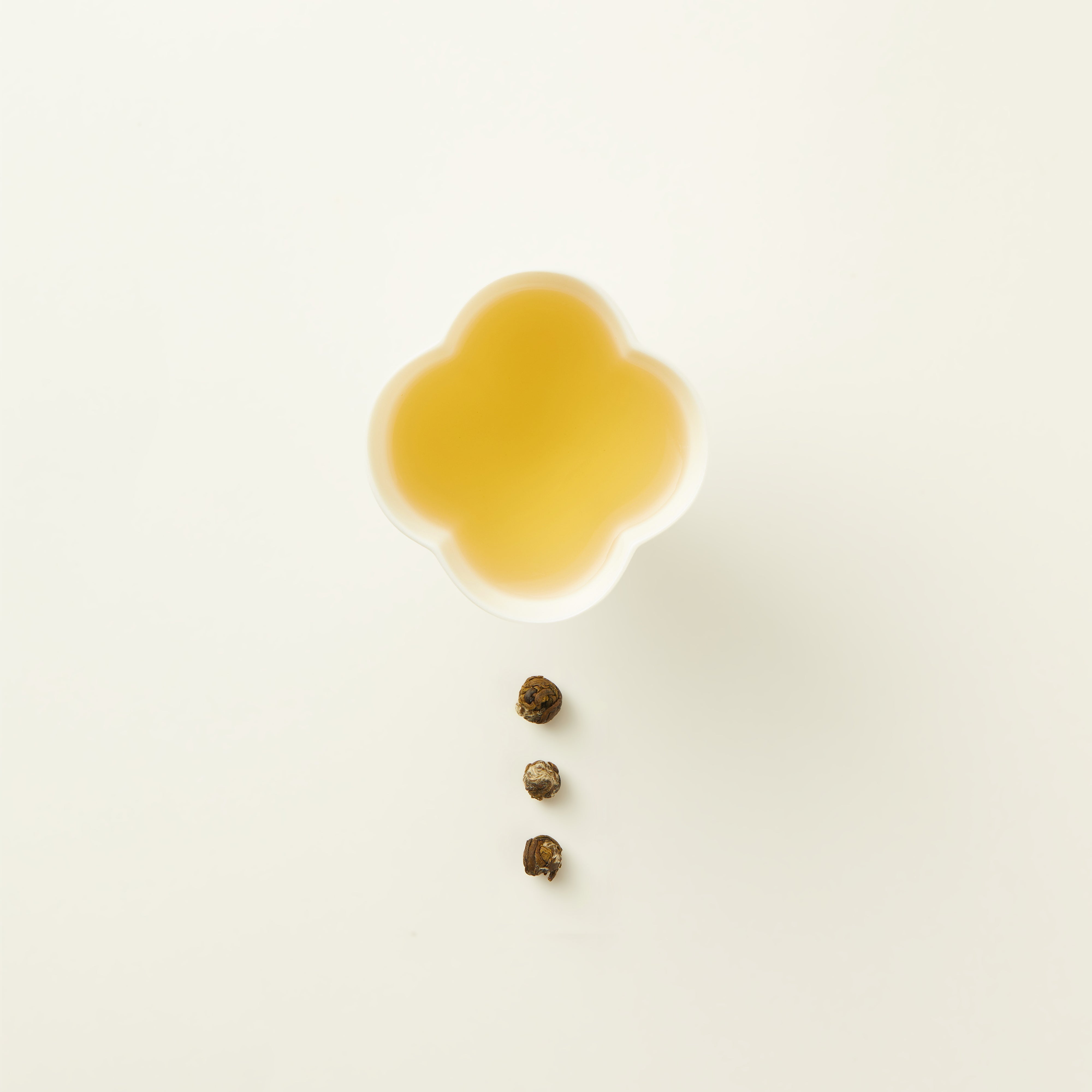 A white clover-shaped cup filled with light yellow tea, placed above three rolled tea leaves arranged in a vertical line on a plain white background.
