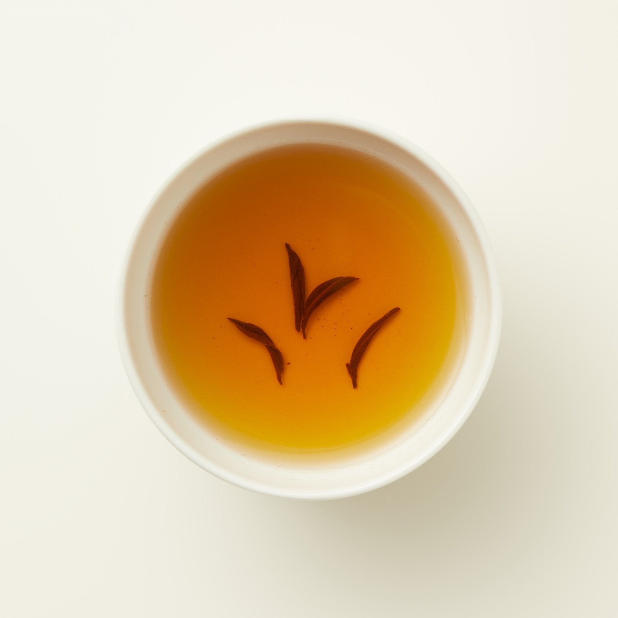 A white cup filled with amber-colored Amber River Black Tea by Chafolio, with golden buds on the surface, viewed from above against a plain background.