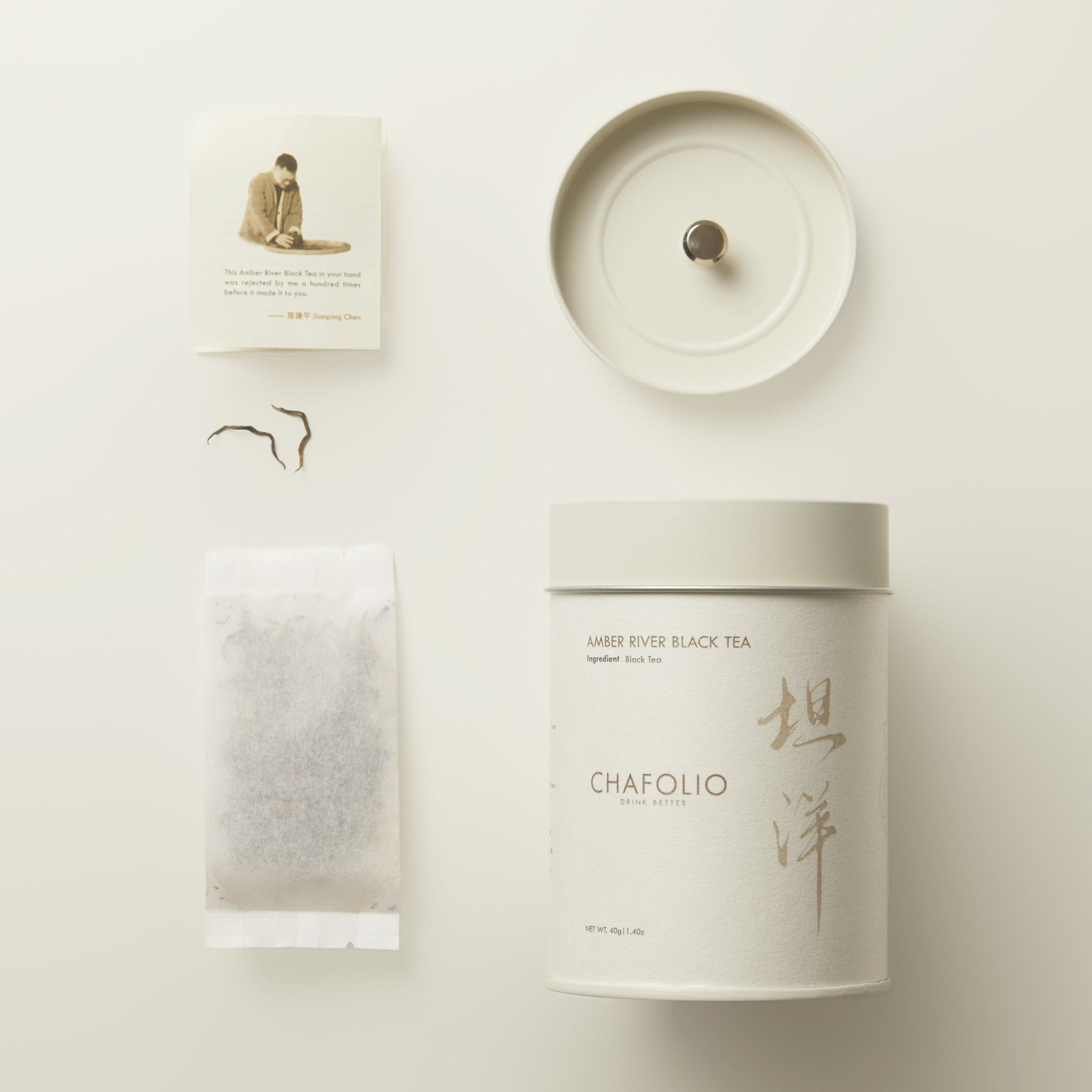 A canister of Chafolio Amber River Black Tea — Craftfolio with its lid, a tea bag, loose golden buds tea leaves, and an info card are displayed neatly on a light surface.