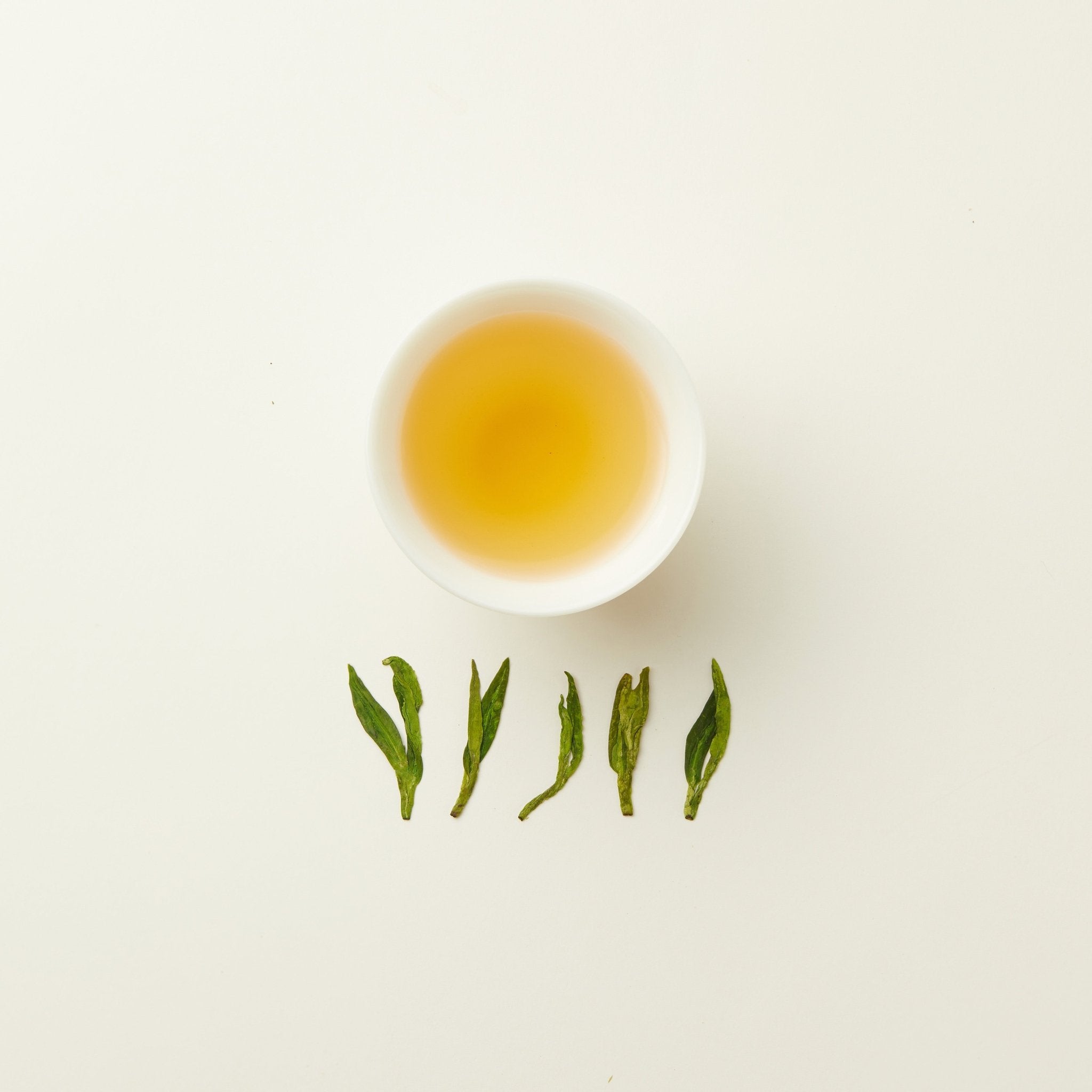 A white cup of light yellow Chafolio Dragon Well tea sits above five green tea leaves on a plain white background.