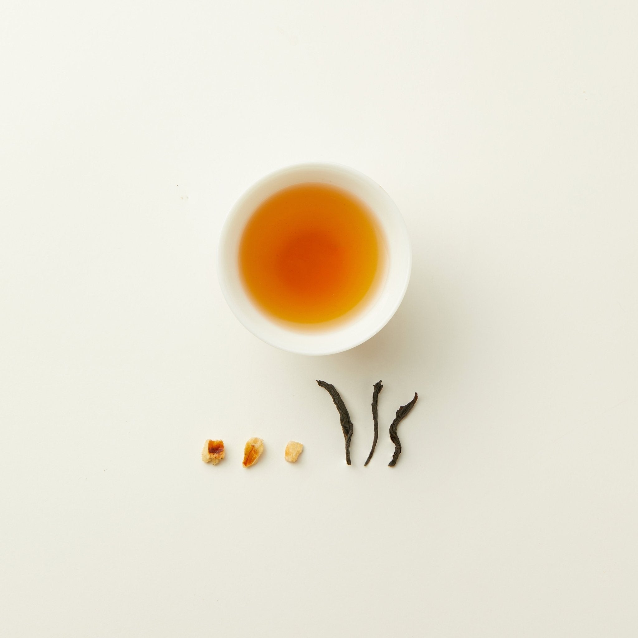 A white cup of Chafolio Lychee Black Tea sits on a white background, with three tea leaves and four dried fruit pieces lined up below it.