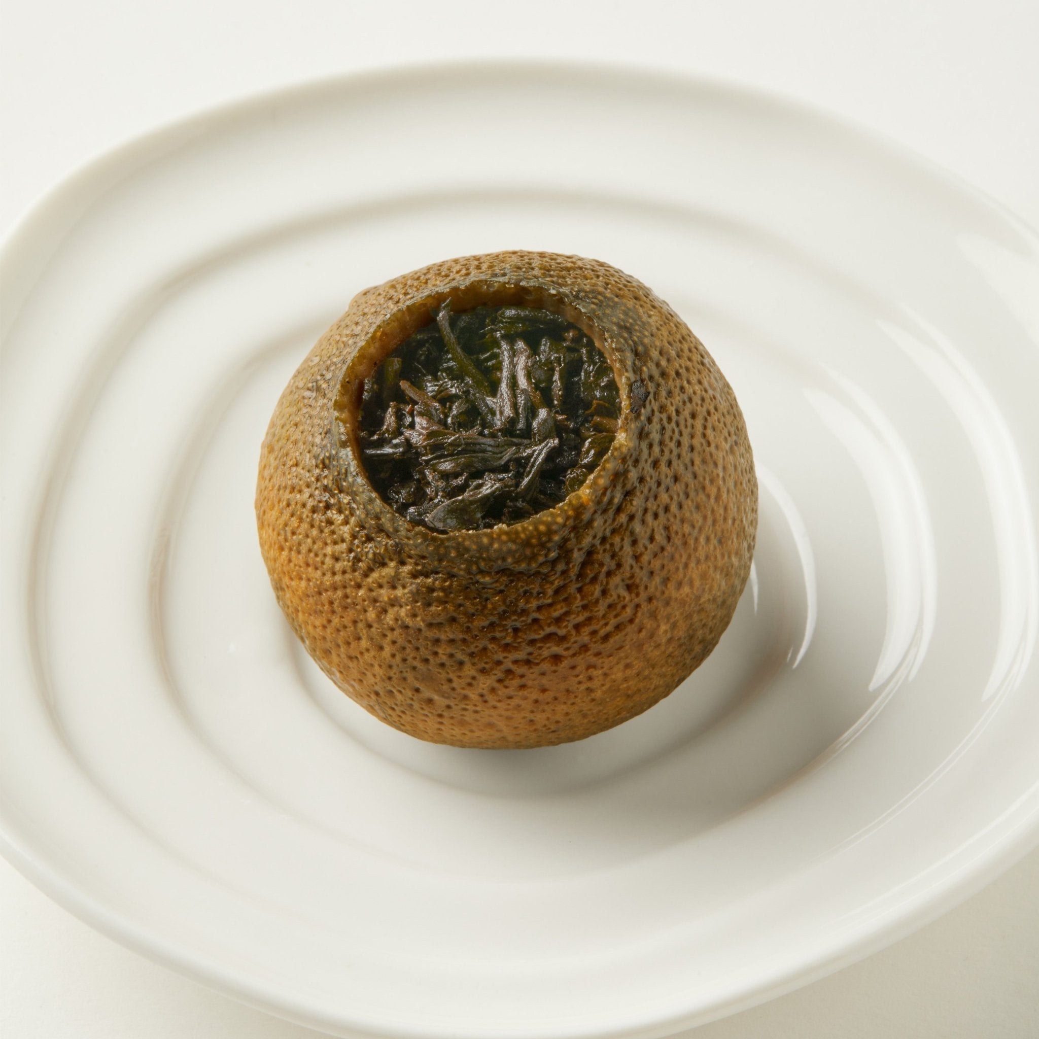 Chafolio’s Tangerine Pu’er, loose leaf pu-er tea in a dried green tangerine peel, is displayed on a white ceramic plate.