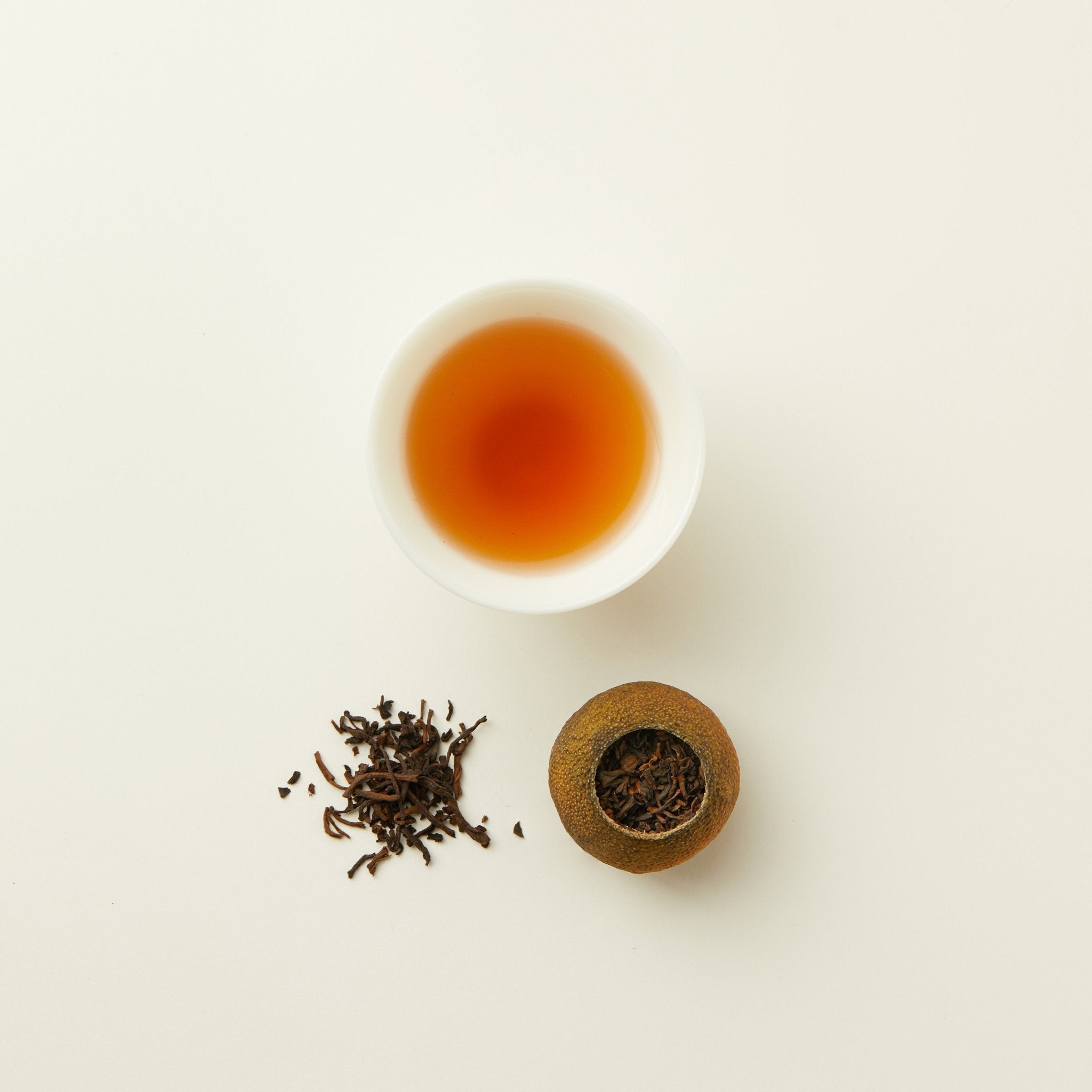 A cup of tea, Chafolio’s Tangerrine Pu'er loose leaf tea, and a dried green tangerine peel filled with tea leaves are arranged on a white surface.