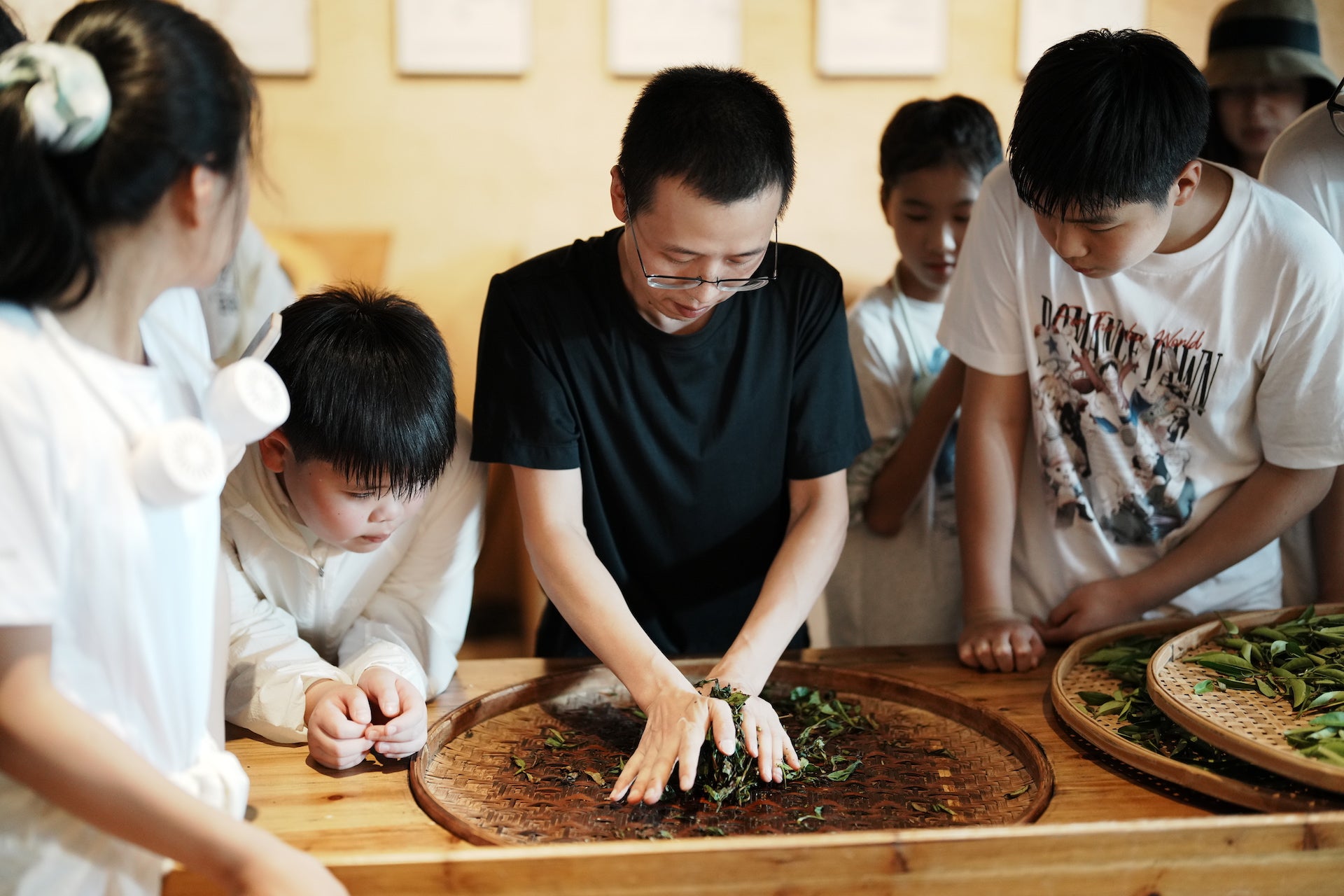 A group of children and an adult gather around a table, with the adult demonstrating how to process tea leaves on a woven tray.