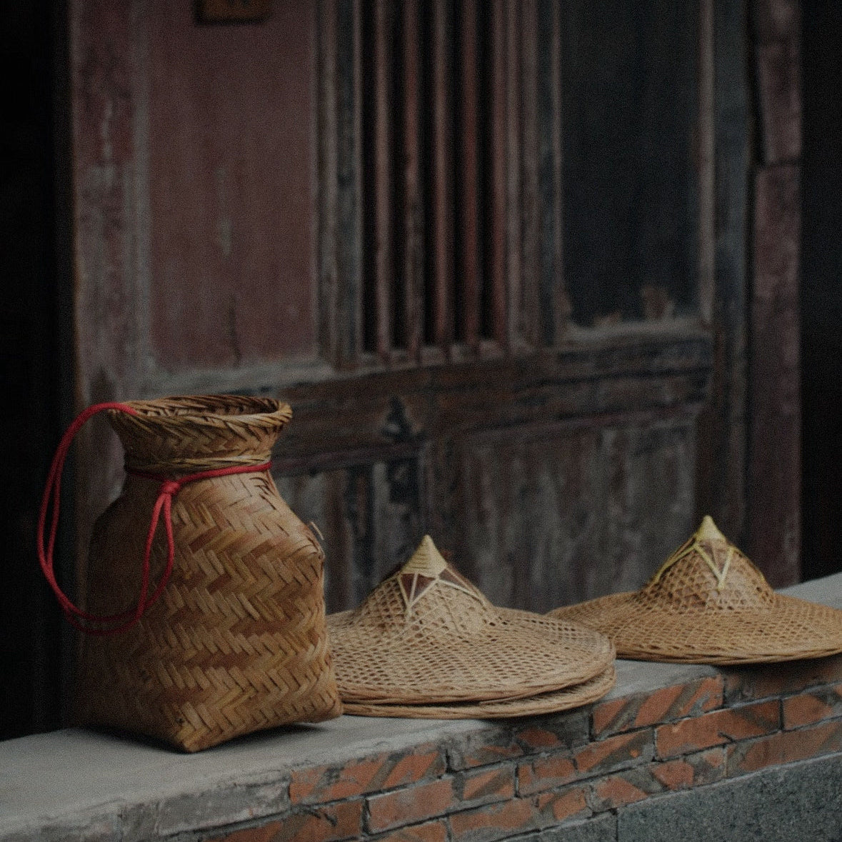 A woven basket with a red handle and two woven hats sit on a brick ledge in front of a weathered wooden door.