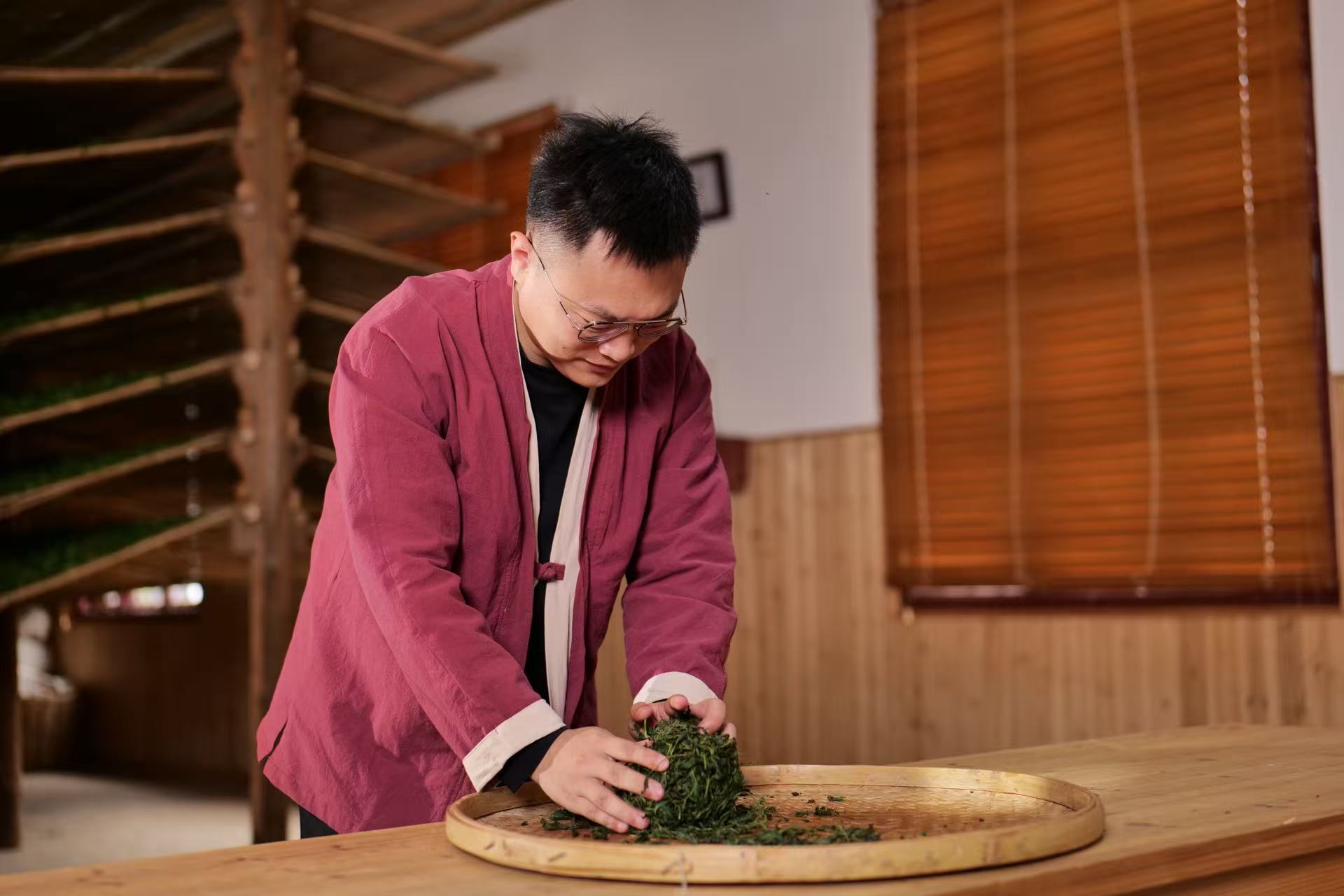A person wearing glasses and a maroon jacket processes tea leaves by hand on a large bamboo tray in a wooden room.