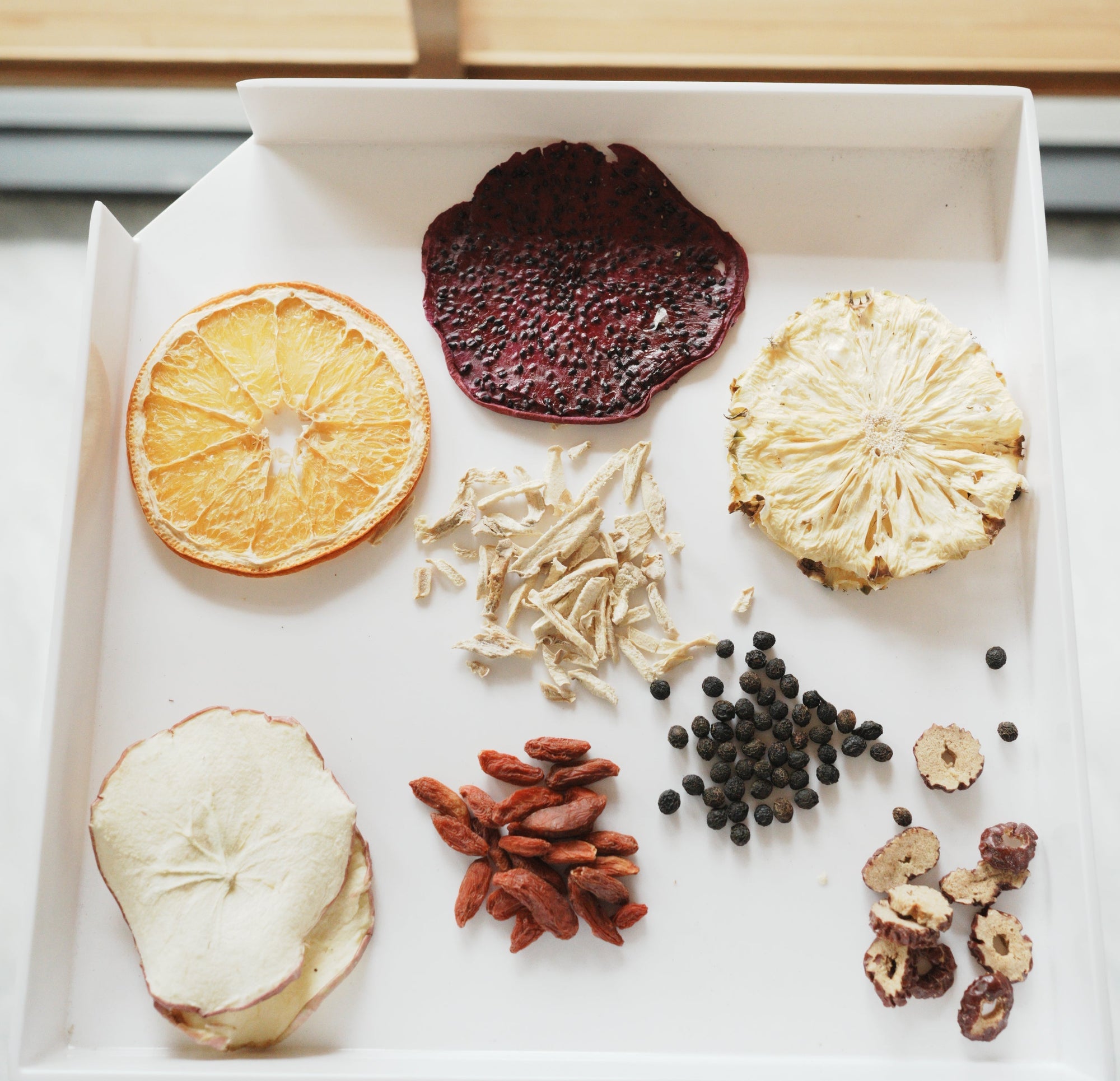 A white tray holds dried apple, orange, pineapple, and dragon fruit slices, goji berries, black peppercorns, dried roots, and hawthorn slices, displayed in separate small piles.
