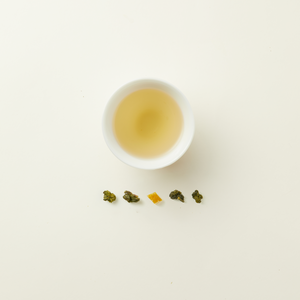 A white cup of Chafolio Yuzu Oolong tea, light yellow in color, is placed above five loose leaf pieces arranged in a line on a plain white background.