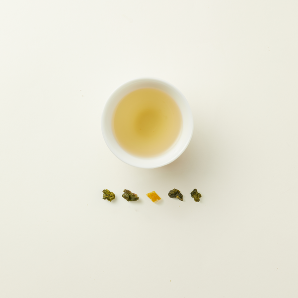 A white cup of Chafolio Yuzu Oolong tea, light yellow in color, is placed above five loose leaf pieces arranged in a line on a plain white background.
