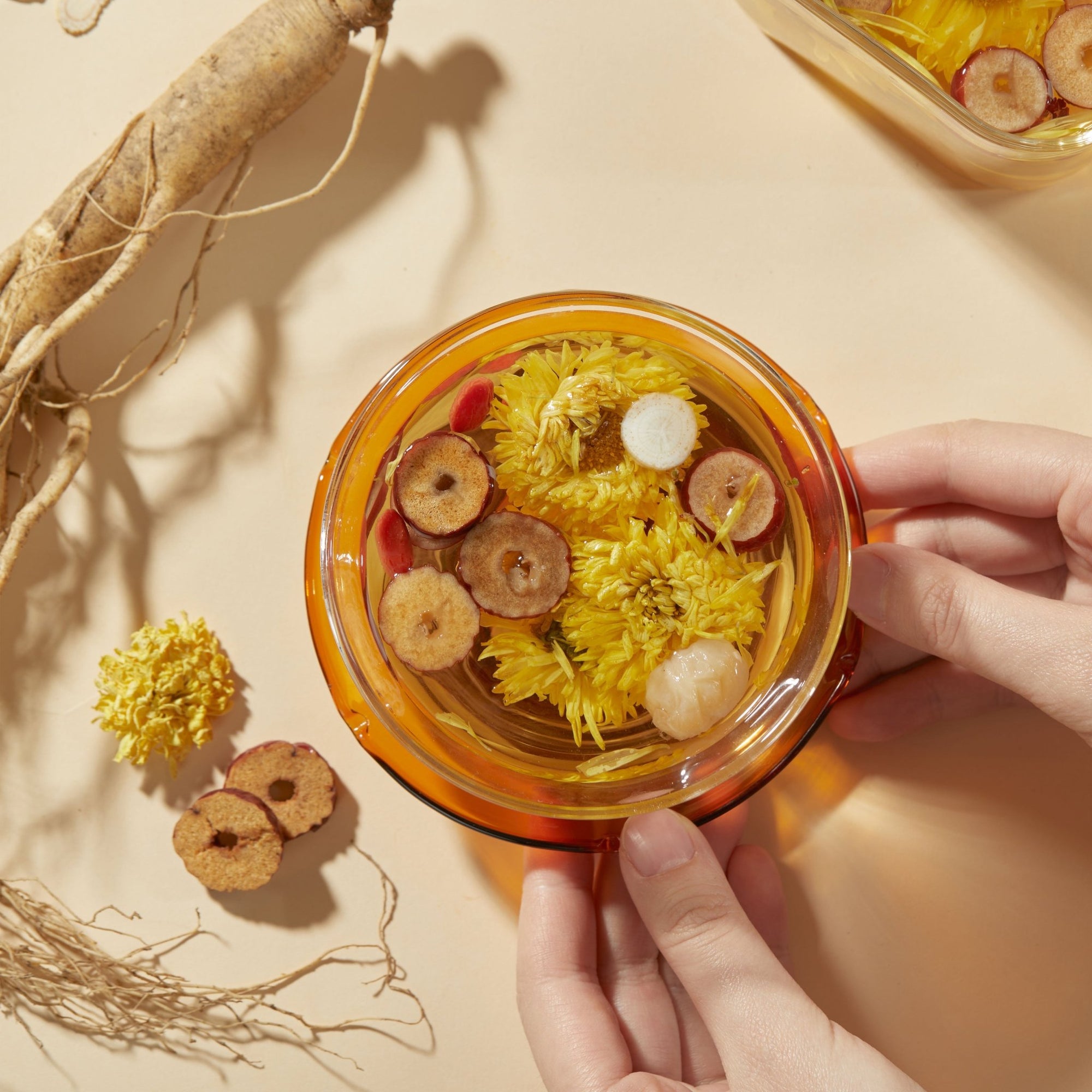 A person holds a glass cup of Chafolio Chrysanthemum Ginseng Tea with yellow flowers, ginseng, and dried fruit slices, while similar traditional Chinese herbal tea ingredients are arranged on the table nearby.