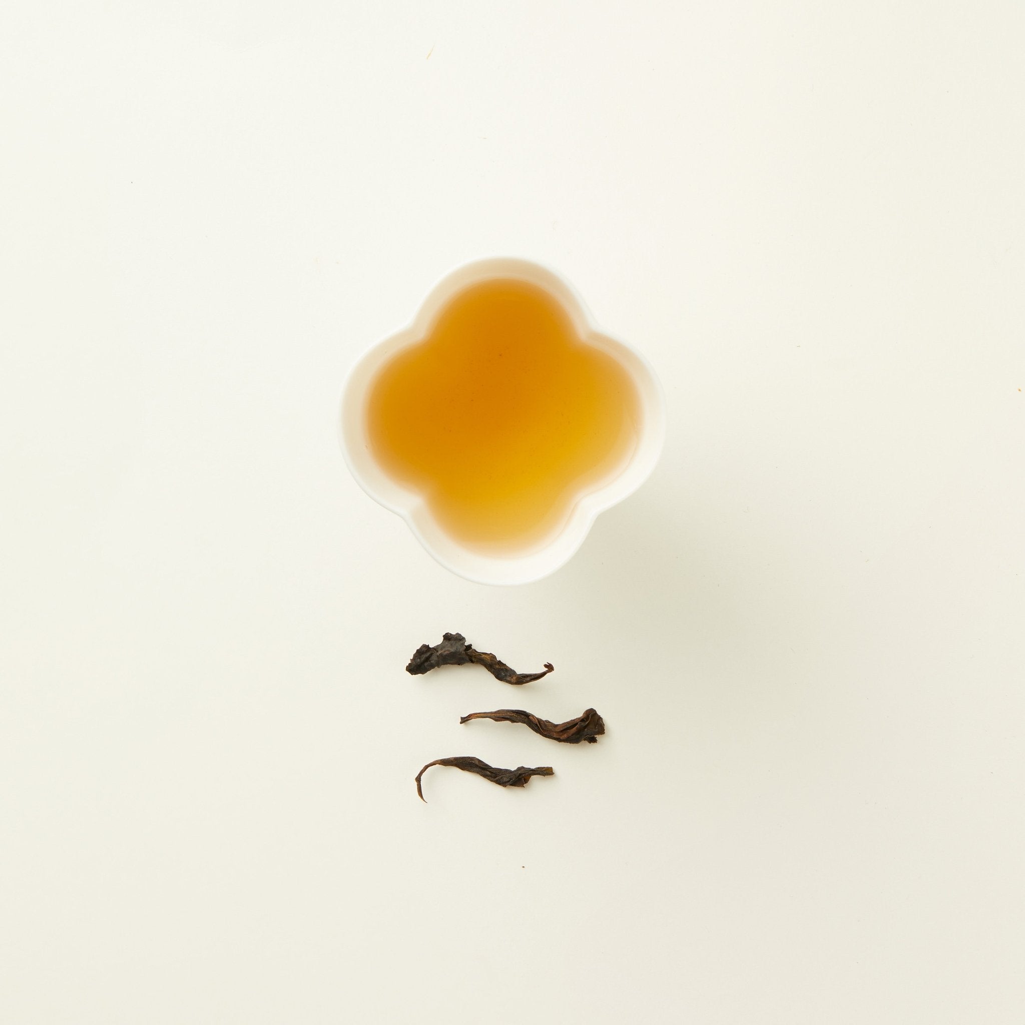 A cup of Chafolio Da Hong Pao — Craftfolio oolong tea sits in a white quatrefoil-shaped cup above three dried tea leaves on a white background.
