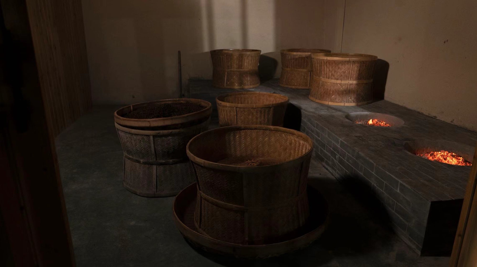 Six large woven baskets are positioned on a dark floor; three sit above built-in brick pits containing glowing embers in a dimly lit room.