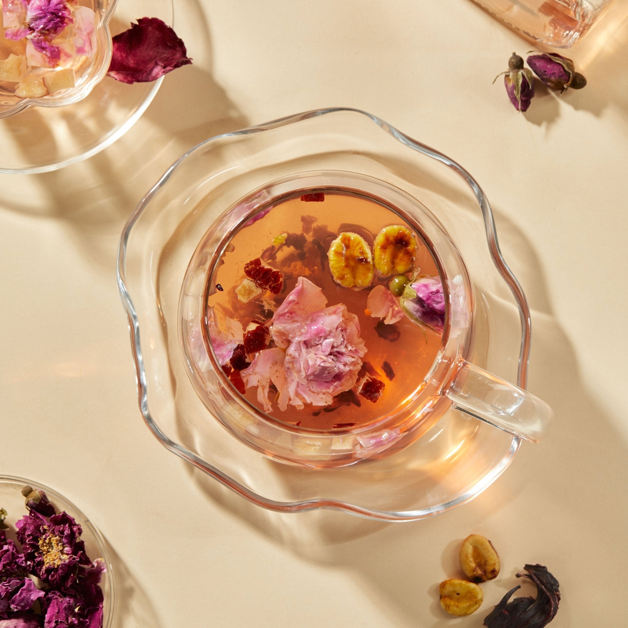 A glass teacup of Chafolio Hibiscus Rose Tea, garnished with dried flowers and monk fruit, sits on a saucer against a beige background with scattered botanicals.