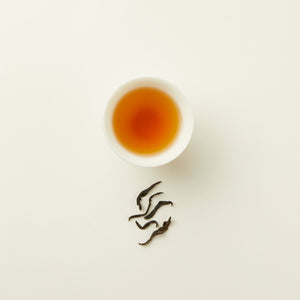 A white cup of amber-colored Chafolio Honey Black Tea sits on a white surface, with a few loose tea leaves arranged beneath it.