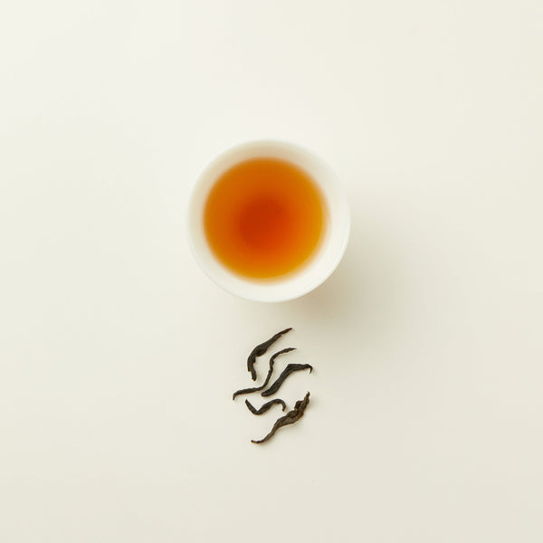 A white cup of amber-colored Chafolio Honey Black Tea sits on a white surface, with a few loose tea leaves arranged beneath it.