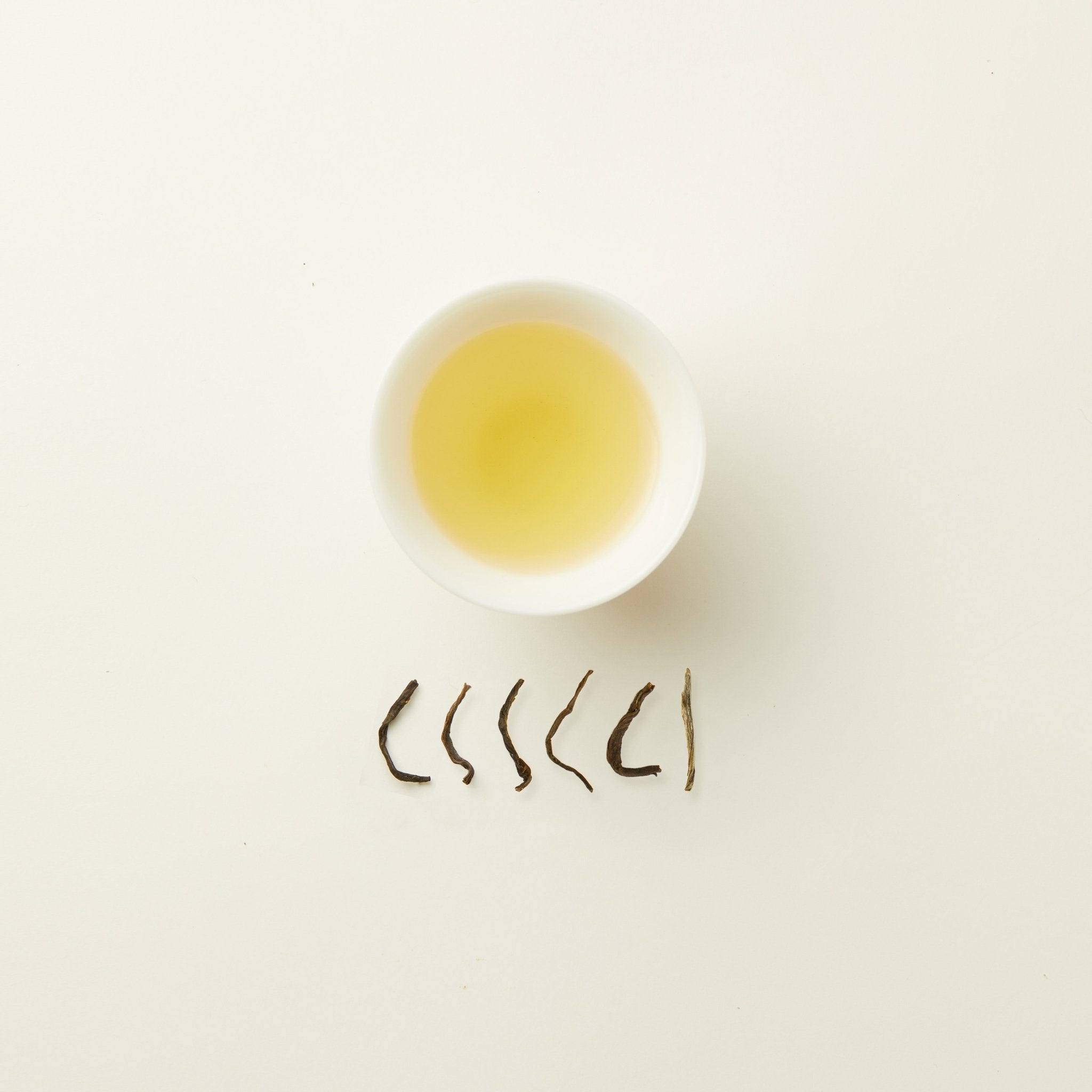 A white cup of light yellow Chafolio Jasmine Green Tea sits above six dried tea leaves arranged in a row on a plain white background.
