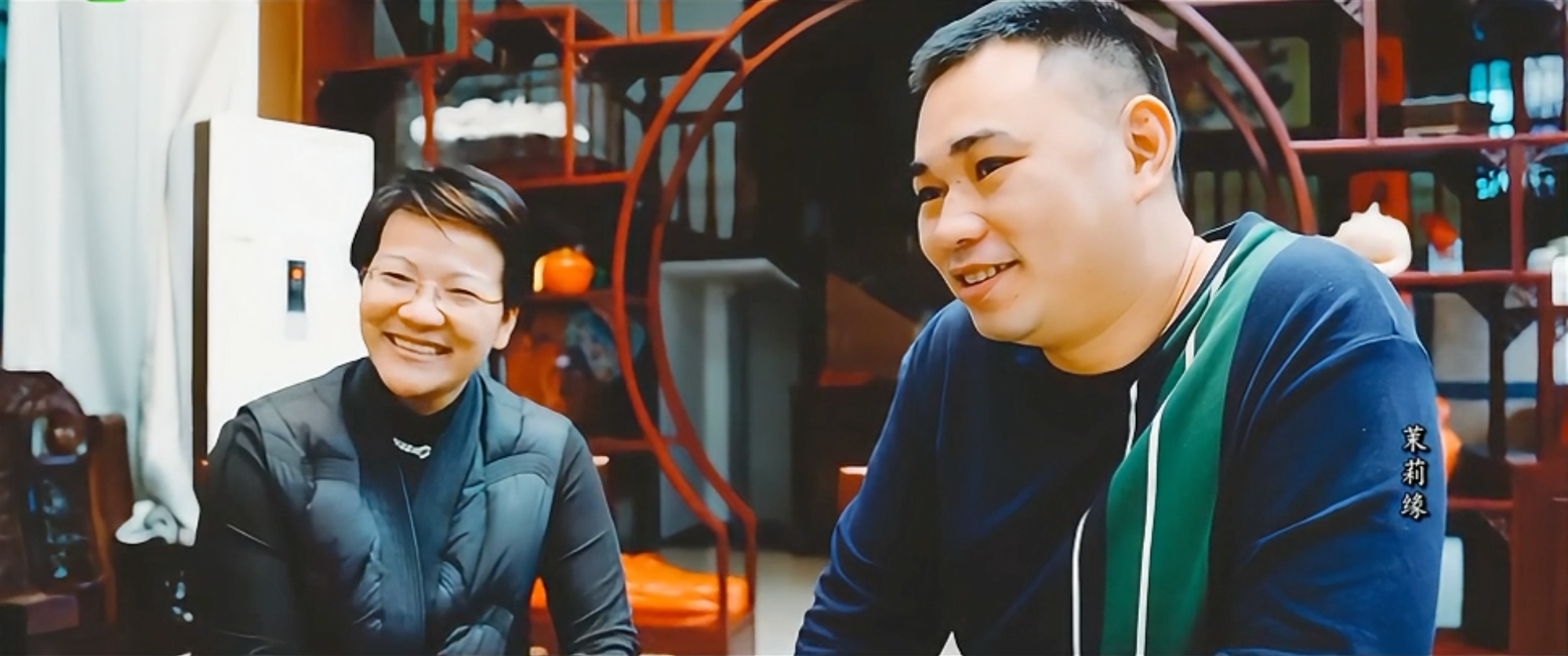 Two people are sitting indoors at a table, smiling and talking. The background includes wooden shelves and a round doorway.
