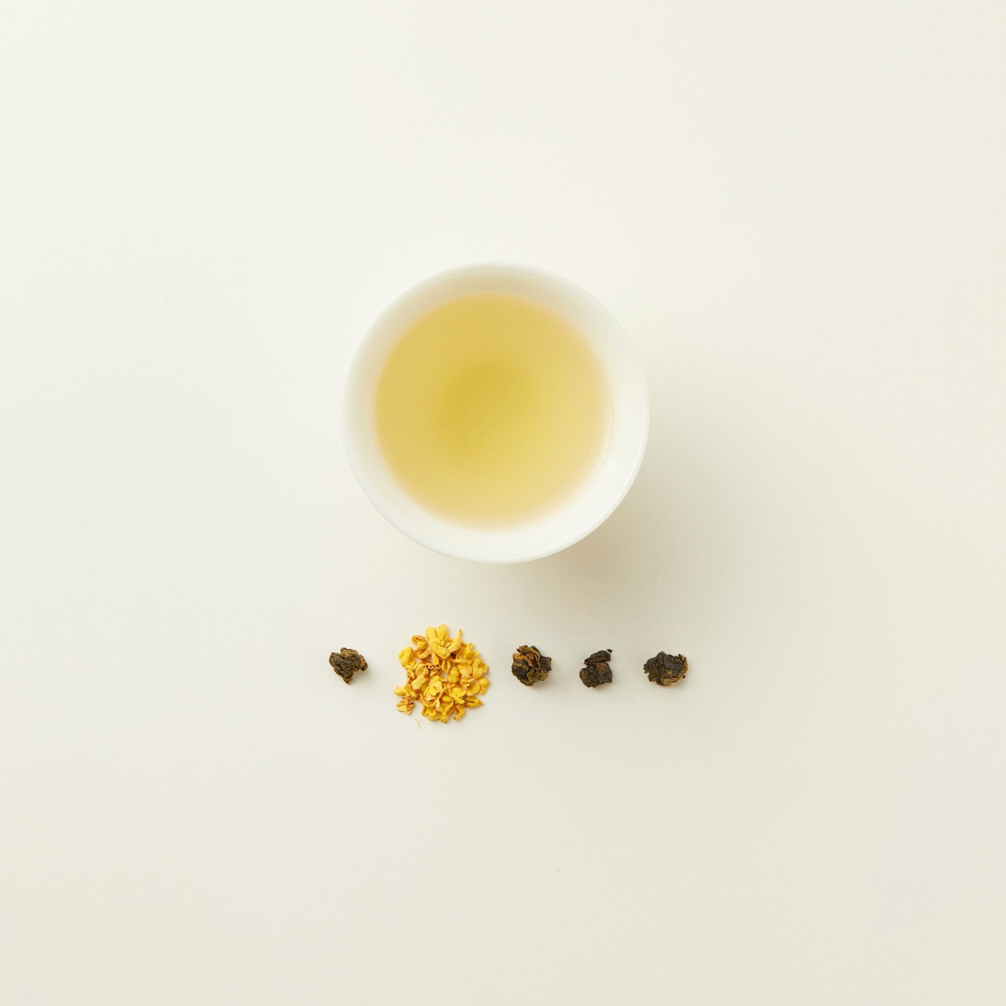 A cup of light yellow Chafolio Osmanthus Oolong tea sits above a row of loose tea leaves and dried yellow flower petals, all arranged on a white background.